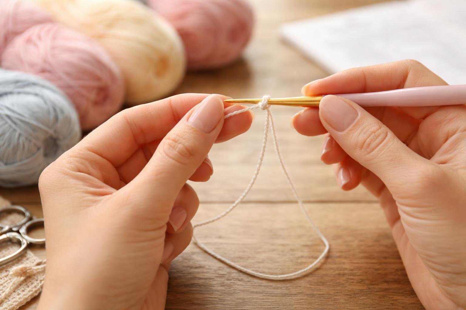 Mãos fazendo uma correntinha de crochê com linha e agulha sobre uma mesa de madeira, com novelos de lã ao redor.